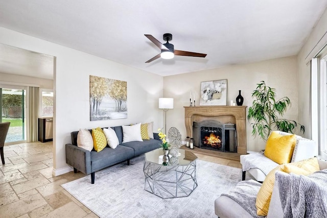 living area featuring a ceiling fan, baseboards, a lit fireplace, and stone tile flooring