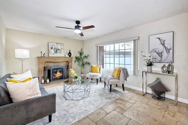 living area featuring a ceiling fan, baseboards, a lit fireplace, and stone tile flooring