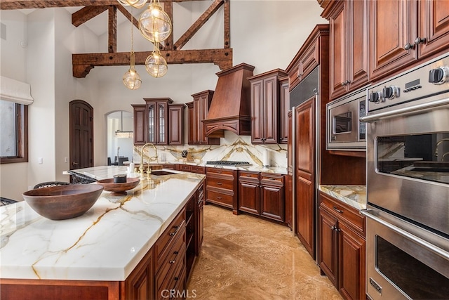 kitchen with a center island with sink, custom range hood, a sink, backsplash, and appliances with stainless steel finishes