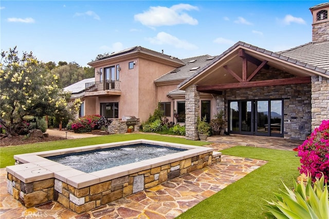 rear view of house featuring stucco siding, a lawn, stone siding, french doors, and a balcony