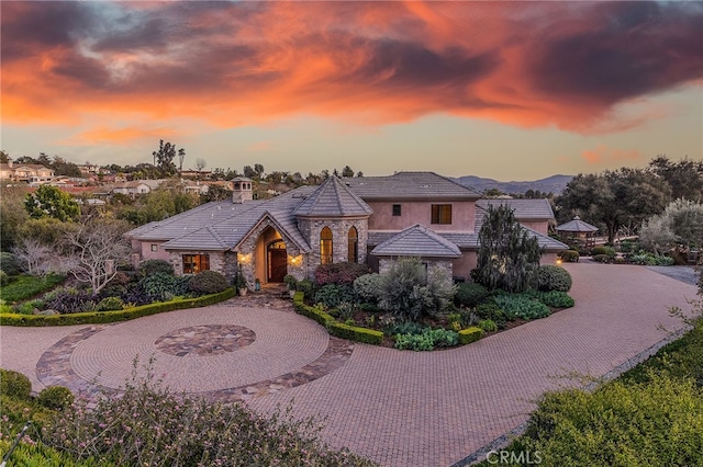 view of front facade featuring stucco siding, a chimney, curved driveway, stone siding, and a tiled roof