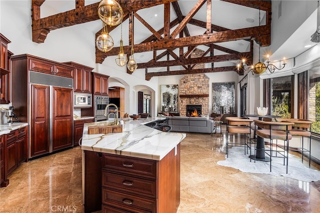 kitchen featuring a center island with sink, reddish brown cabinets, arched walkways, a stone fireplace, and built in appliances