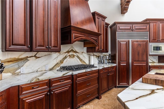 kitchen with light stone counters, decorative backsplash, custom range hood, built in appliances, and reddish brown cabinets