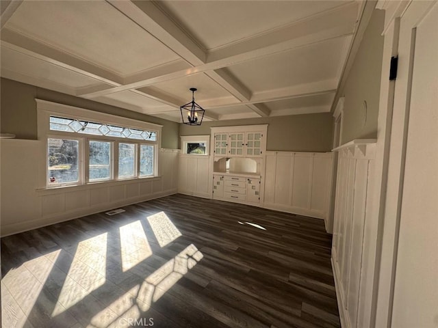 empty room featuring beamed ceiling, dark wood-type flooring, coffered ceiling, an inviting chandelier, and wainscoting