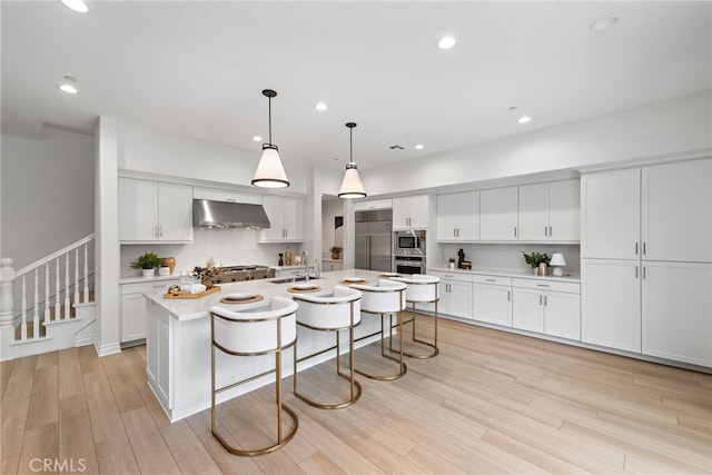 kitchen featuring an island with sink, light countertops, light wood-style floors, built in appliances, and under cabinet range hood