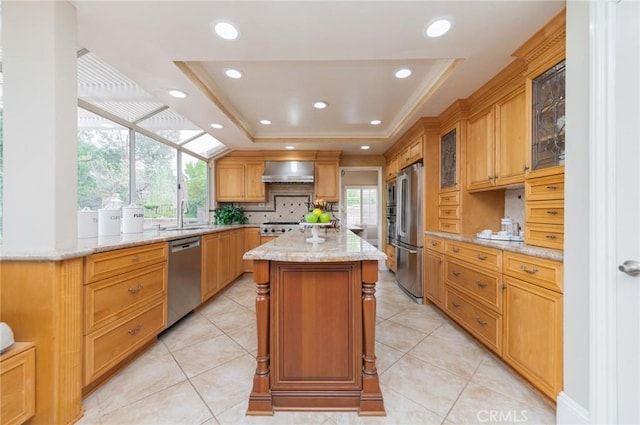 kitchen with a center island, stainless steel appliances, wall chimney exhaust hood, a raised ceiling, and light stone countertops