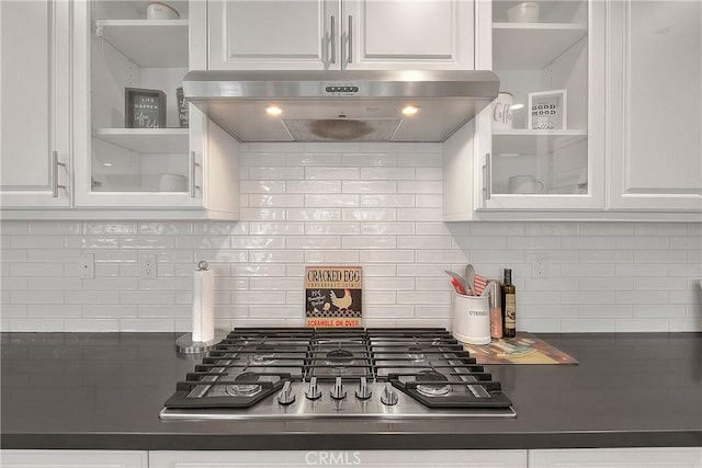 kitchen with under cabinet range hood, dark countertops, white cabinetry, stainless steel gas stovetop, and glass insert cabinets