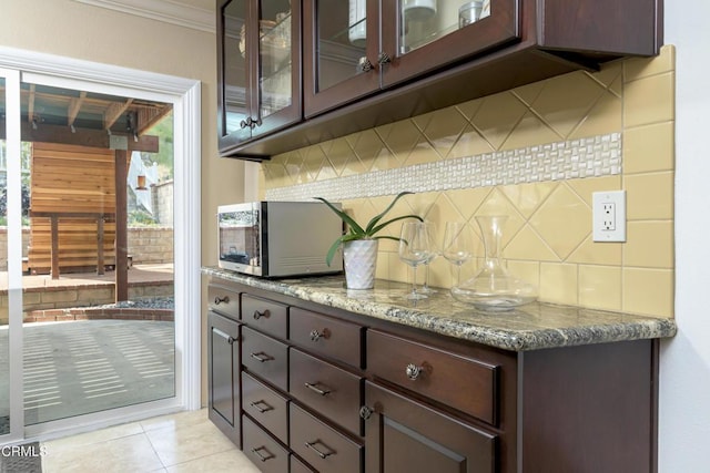 kitchen with tasteful backsplash, stainless steel microwave, dark brown cabinets, glass insert cabinets, and light tile patterned floors