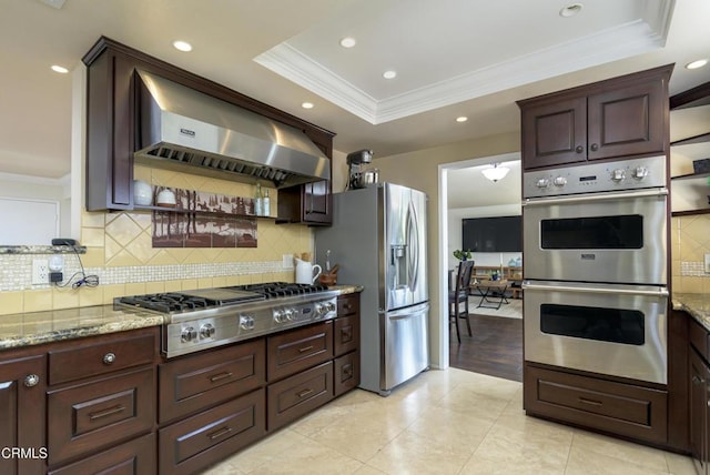 kitchen featuring light stone countertops, ornamental molding, exhaust hood, stainless steel appliances, and a raised ceiling