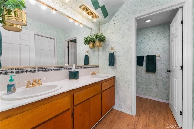 full bathroom featuring a sink, wood finished floors, double vanity, and wallpapered walls