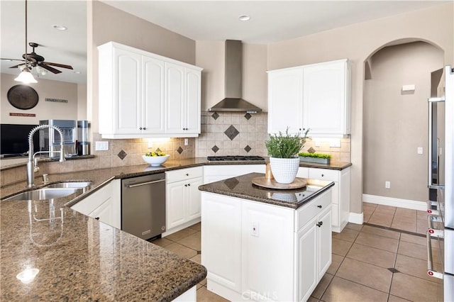 kitchen with a ceiling fan, a sink, dark stone countertops, stainless steel appliances, and wall chimney range hood