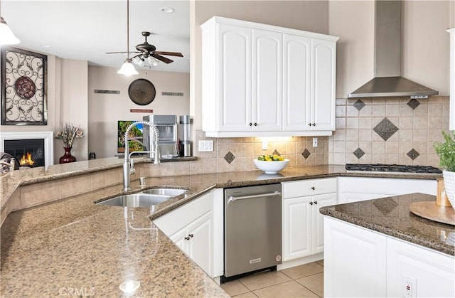 kitchen featuring wall chimney range hood, stainless steel gas cooktop, dark stone counters, a ceiling fan, and a sink