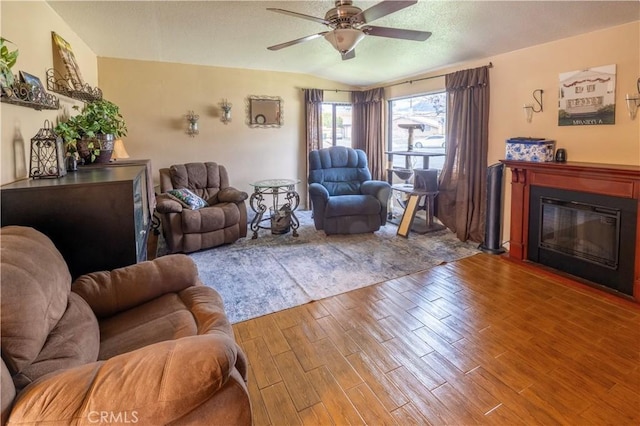 living area featuring ceiling fan, vaulted ceiling, wood finished floors, a glass covered fireplace, and a textured ceiling