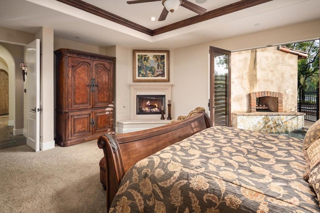 bedroom with crown molding, light colored carpet, a tray ceiling, a warm lit fireplace, and arched walkways