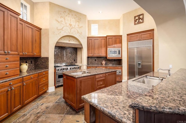 kitchen featuring built in appliances, an island with sink, dark stone countertops, brown cabinets, and a sink