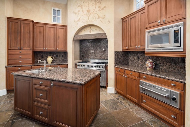 kitchen with brown cabinetry, an island with sink, a sink, appliances with stainless steel finishes, and backsplash