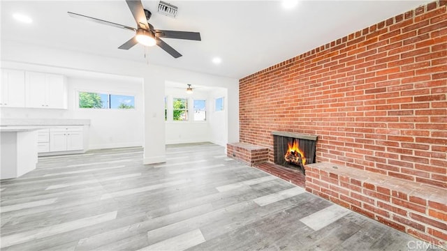 unfurnished living room featuring visible vents, brick wall, light wood-style flooring, a fireplace, and ceiling fan