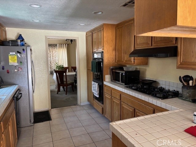 kitchen featuring visible vents, tile counters, black appliances, under cabinet range hood, and brown cabinets