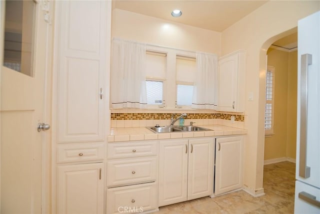 kitchen with arched walkways, a sink, tile counters, white cabinetry, and backsplash