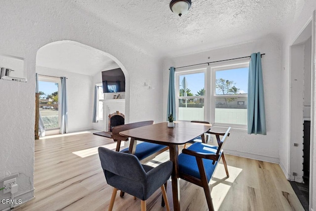 dining space featuring a fireplace, light wood-style floors, a textured wall, arched walkways, and a textured ceiling