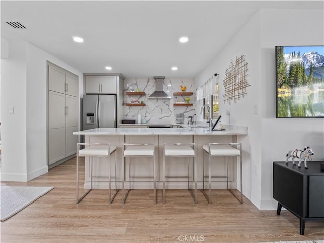 kitchen with a peninsula, wall chimney exhaust hood, stainless steel fridge with ice dispenser, light countertops, and decorative backsplash