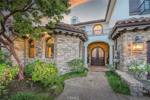 exterior entry at dusk featuring a tile roof, stucco siding, french doors, a balcony, and stone siding