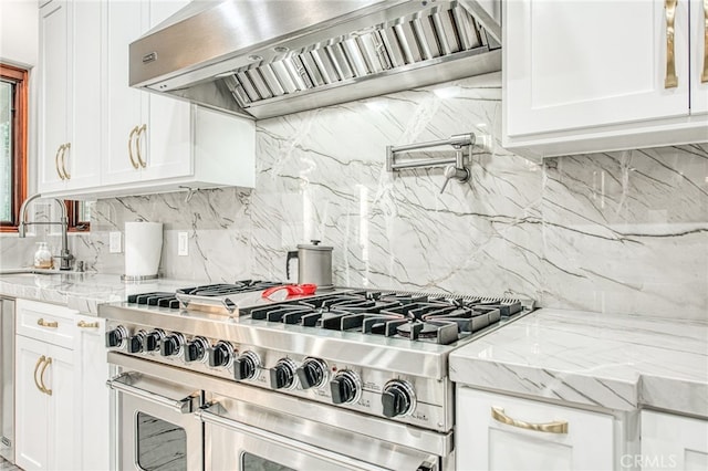 kitchen with a sink, double oven range, light stone counters, wall chimney exhaust hood, and white cabinets