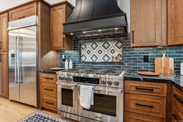 kitchen with brown cabinetry, light wood-type flooring, custom exhaust hood, high quality appliances, and tasteful backsplash