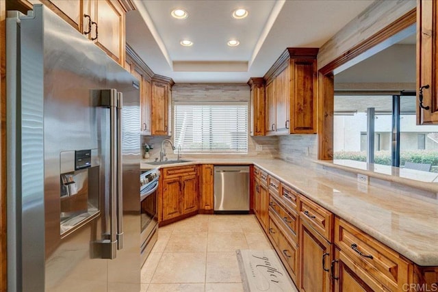 kitchen featuring a tray ceiling, decorative backsplash, appliances with stainless steel finishes, brown cabinetry, and a sink