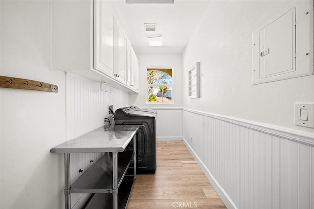 clothes washing area featuring a wainscoted wall, washing machine and clothes dryer, electric panel, cabinet space, and light wood-type flooring