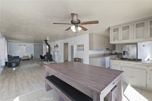 kitchen with a ceiling fan, appliances with stainless steel finishes, light countertops, glass insert cabinets, and a wood stove