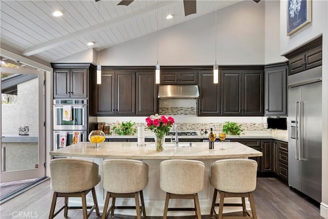 kitchen featuring an island with sink, under cabinet range hood, backsplash, appliances with stainless steel finishes, and dark wood-style flooring