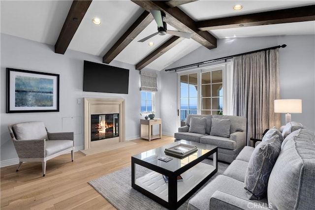 living room featuring vaulted ceiling with beams, baseboards, ceiling fan, light wood-type flooring, and a glass covered fireplace