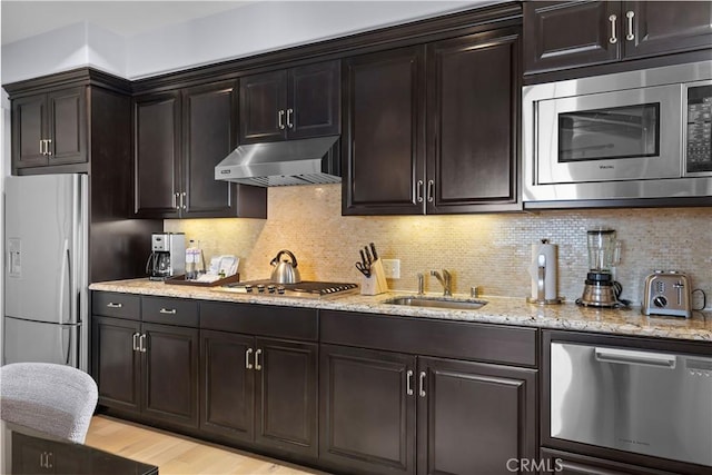 kitchen with tasteful backsplash, under cabinet range hood, light stone counters, appliances with stainless steel finishes, and a sink