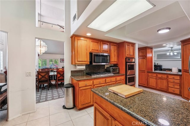 kitchen with dark stone countertops, ceiling fan with notable chandelier, brown cabinets, and stainless steel appliances