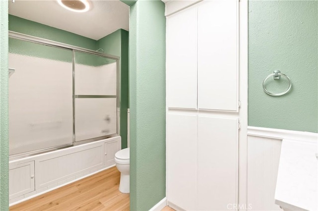 bathroom with toilet, wood finished floors, shower / bath combination with glass door, and wainscoting