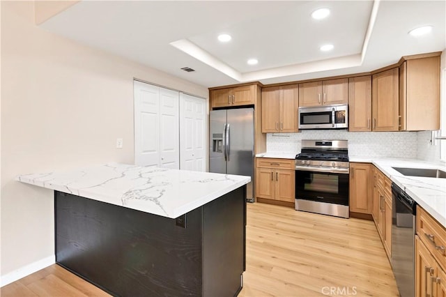 kitchen with a tray ceiling, appliances with stainless steel finishes, light wood finished floors, decorative backsplash, and light stone countertops