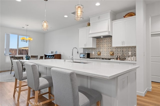 kitchen featuring an island with sink, custom range hood, tasteful backsplash, light wood finished floors, and light countertops