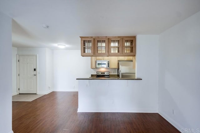 kitchen with a peninsula, dark wood-style flooring, stainless steel appliances, glass insert cabinets, and dark countertops