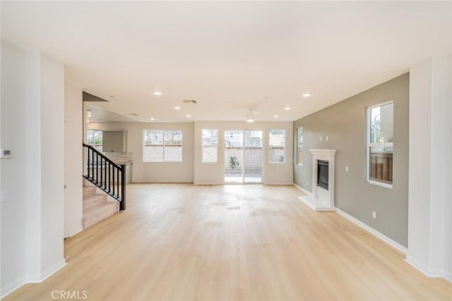 unfurnished living room featuring baseboards, stairs, light wood-type flooring, a glass covered fireplace, and a ceiling fan