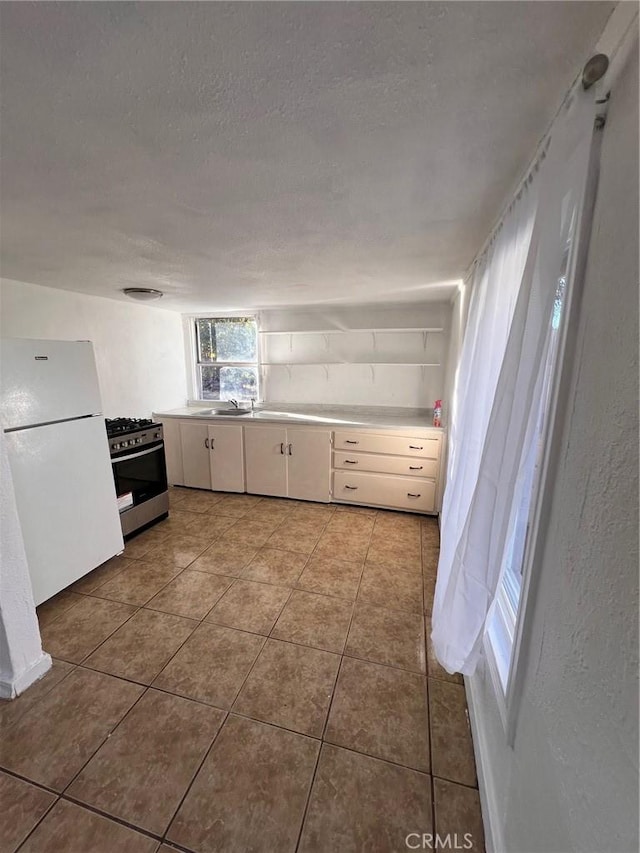 kitchen featuring freestanding refrigerator, white cabinets, a textured ceiling, tile patterned floors, and stainless steel gas range oven