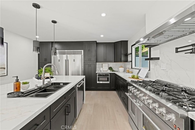 kitchen featuring a sink, appliances with stainless steel finishes, wall chimney exhaust hood, light stone countertops, and dark cabinets