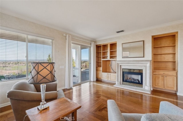 living room with built in shelves, wood finished floors, visible vents, ornamental molding, and a glass covered fireplace