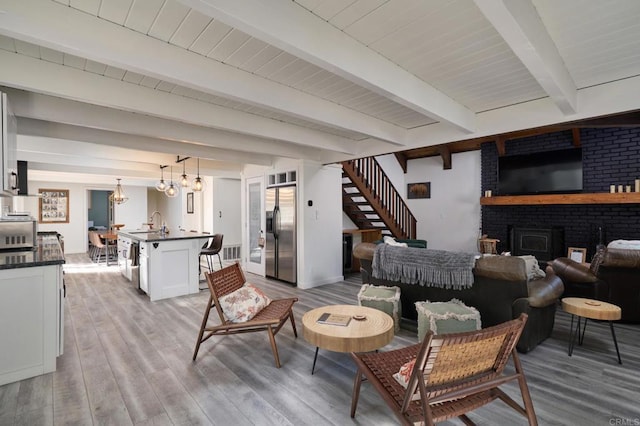 living area with wood finished floors, beam ceiling, stairs, a brick fireplace, and a notable chandelier