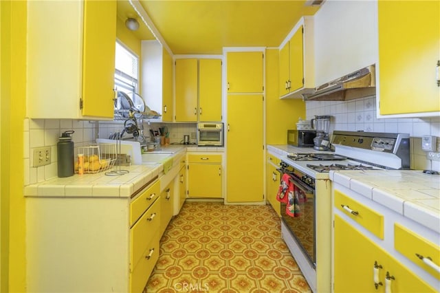 kitchen featuring white gas stove, decorative backsplash, tile countertops, and under cabinet range hood