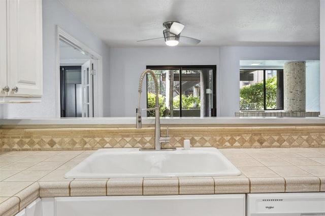 interior space with ceiling fan, tile countertops, dishwashing machine, white cabinetry, and a sink