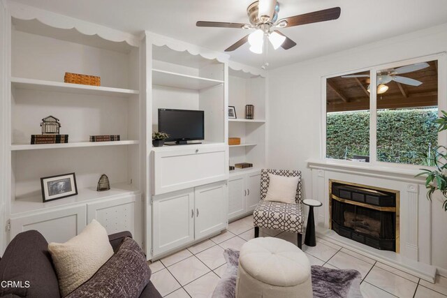 living area with a glass covered fireplace, built in shelves, ceiling fan, and light tile patterned floors