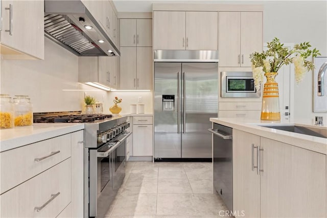 kitchen with light countertops, wall chimney range hood, built in appliances, and a sink