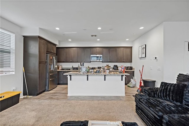 kitchen featuring light stone counters, a kitchen island with sink, stainless steel appliances, dark brown cabinetry, and a kitchen bar