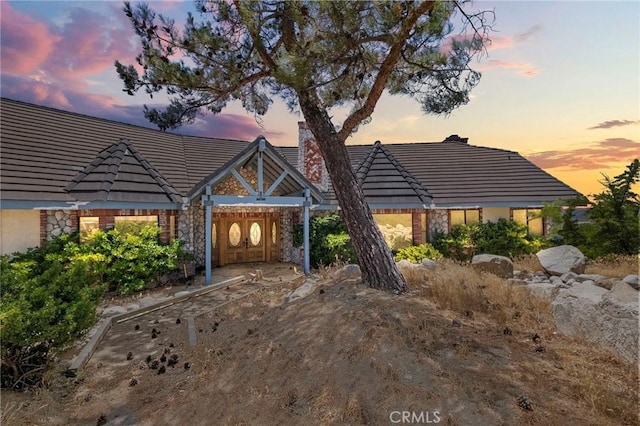 back of house at dusk with a tiled roof, stone siding, and a chimney